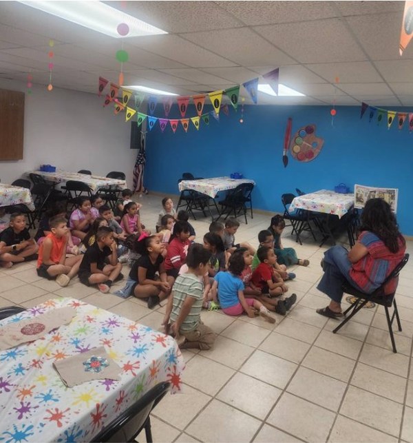 Summer Reading - Van Horn.jpg Children seated on the floor listening to a librarian read a story in a decorated library room.